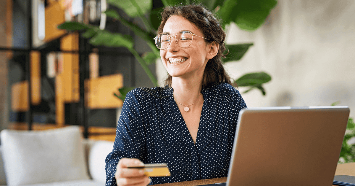 Woman in glasses at laptop, smiling with a debit card in one hand