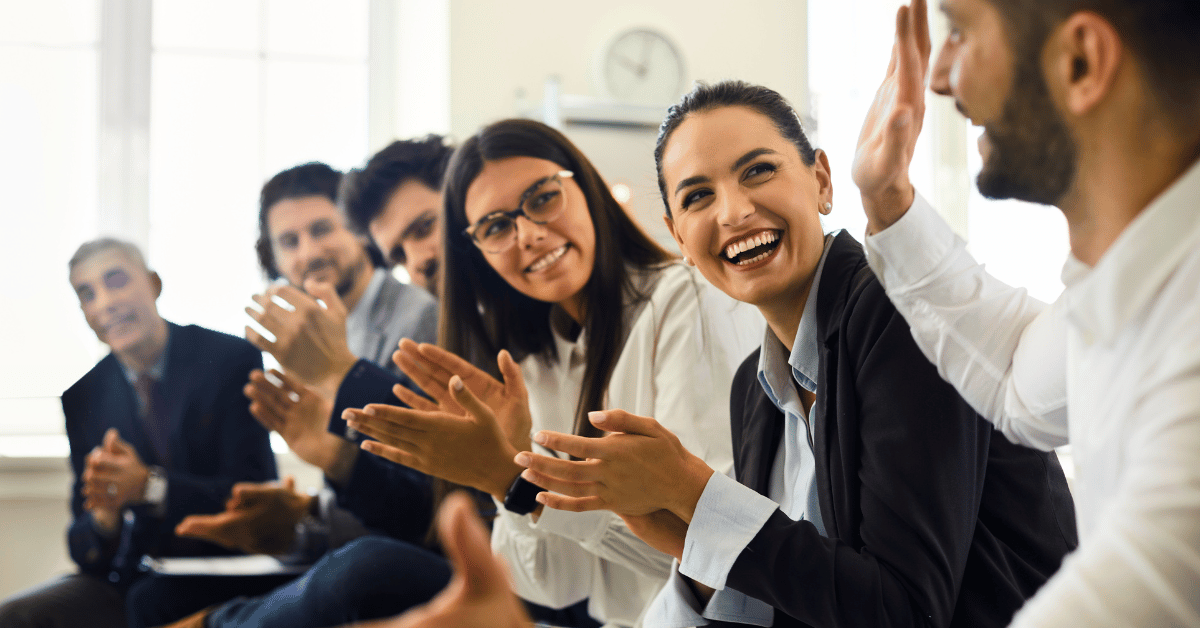 Five employees on the left clap while one employee on the right raises his hand - representing collaboration and success involved with sales incentive program examples