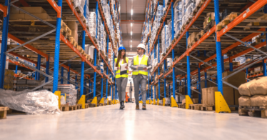 Two people, a man and woman, walking through a distribution warehouse wearing hard hats.