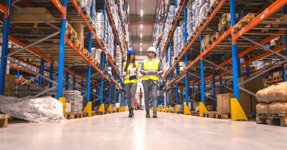 Two people, a man and woman, walking through a distribution warehouse wearing hard hats.